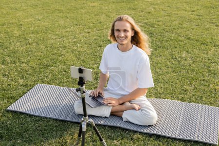 carefree yoga man looking at camera while sitting with laptop near tripod with mobile phone on green lawn