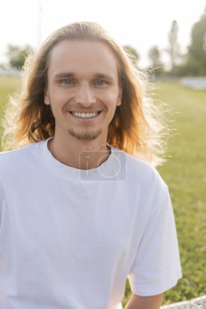 portrait of overjoyed long haired man in white cotton t-shirt smiling at camera outdoors