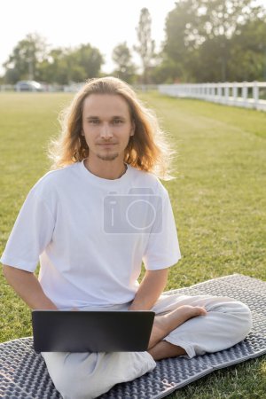 positif homme de yoga aux cheveux longs assis dans une pose facile près d'un ordinateur portable et regardant la caméra sur la pelouse verte