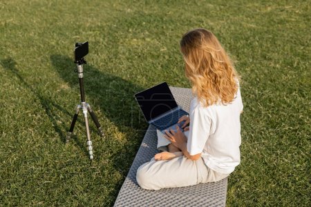 Téléchargez les photos : Vue grand angle de l'homme aux cheveux longs en utilisant un ordinateur portable près du trépied avec téléphone portable pendant la leçon de yoga sur le champ vert - en image libre de droit