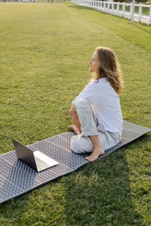 Langhaariger Mann sitzt in Salbeipose neben Laptop und schaut auf Yogamatte auf der grünen Wiese weg