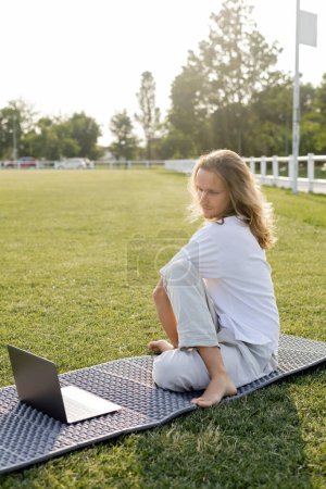 Langhaariger Mann sitzt in Salbeipose während Online-Unterricht auf Laptop auf grünem Rasen des Freiluftstadions