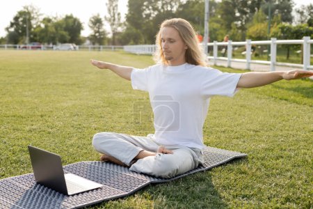 Téléchargez les photos : Jeune homme assis dans la pose facile avec les mains tendues pendant la leçon de yoga en ligne sur ordinateur portable à l'extérieur - en image libre de droit