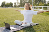 young man sitting in easy pose with outstretched hands during online yoga lesson on laptop outdoors puzzle #648520014