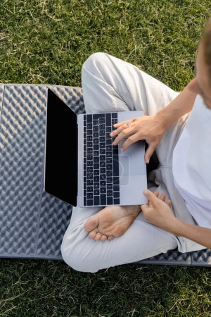 Téléchargez les photos : Vue du haut de l'homme cultivé en utilisant un ordinateur portable tout en étant assis dans la pose de yoga facile sur pelouse herbeuse - en image libre de droit