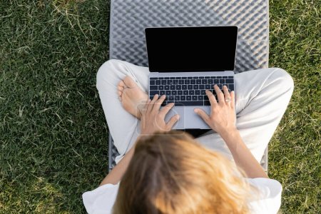 Draufsicht des barfüßigen Mannes, der in lockerer Pose sitzt und Laptop auf Yogamatte auf grünem Rasen benutzt