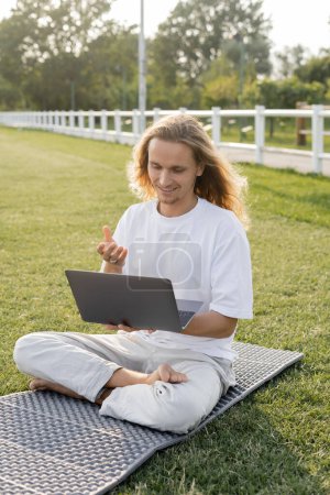 smiling man talking during yoga lesson on laptop while sitting in easy pose on outdoor stadium