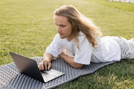 junger langhaariger Mann schaut bei Yoga-Stunde auf der grünen Wiese auf Laptop