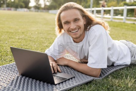 young and carefree man looking at camera while lying near laptop on yoga mat 