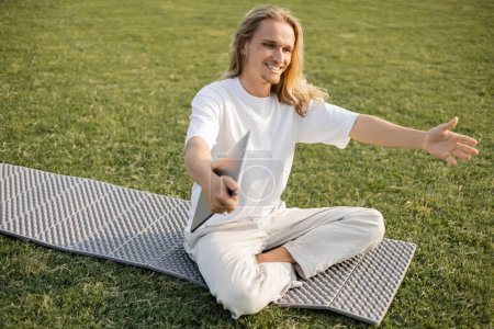 Überglücklicher Mann mit Laptop zeigt Grußgeste beim Sitzen auf Yogamatte auf grünem Rasen im Freien