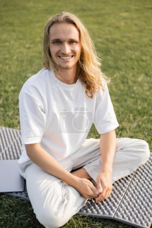 hombre despreocupado sentado en la esterilla de yoga cerca de la computadora portátil y sonriendo a la cámara al aire libre