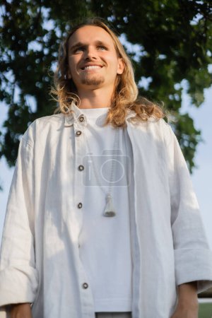 low angle view of carefree long haired yoga man in linen clothes smiling outdoors