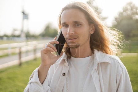 elegante hombre de yoga de pelo largo mirando a la cámara durante la conversación en el teléfono móvil al aire libre