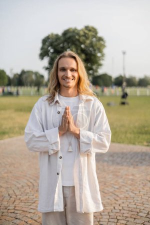 long haired yoga man looking at camera and showing anjali mudra gesture while standing outdoors