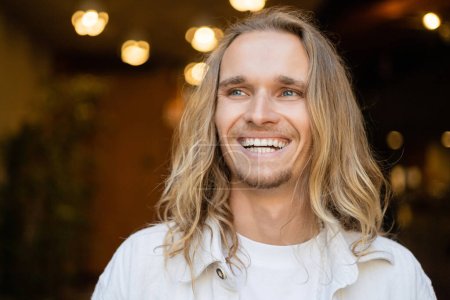 Téléchargez les photos : Portrait de joyeux homme de yoga avec de longs cheveux blonds regardant loin près des lumières floues de la ville sur le fond - en image libre de droit