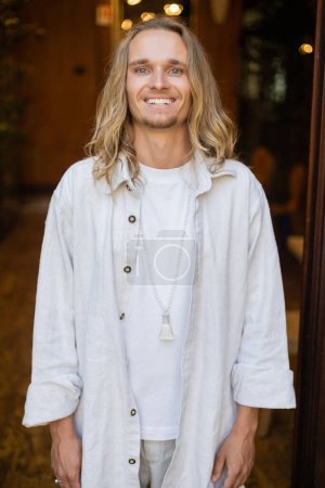 happy long haired yoga man in white cotton clothes and beads smiling at camera outdoors