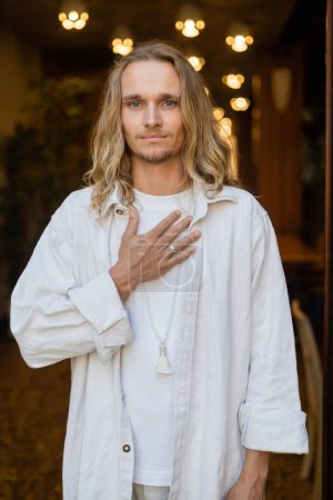 Photo for Fair haired yoga man in white linen clothes touching chest and looking at camera near city lights on blurred background - Royalty Free Image