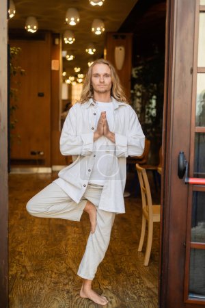 Photo for Full length of barefoot man in white linen clothes standing in tree pose with praying hands in house with blurred lights - Royalty Free Image