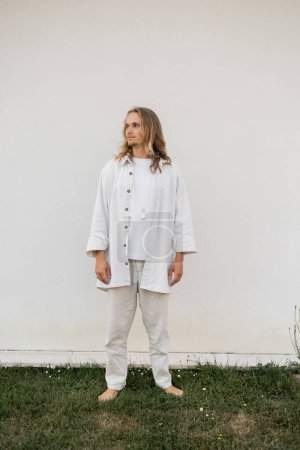 Foto de Longitud completa de hombre joven descalzo en ropa de lino blanco de pie cerca de la pared al aire libre y mirando hacia otro lado - Imagen libre de derechos