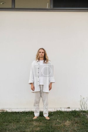 Téléchargez les photos : Pleine longueur de pieds nus et homme aux cheveux blonds dans des vêtements en lin debout près du mur blanc à l'extérieur - en image libre de droit