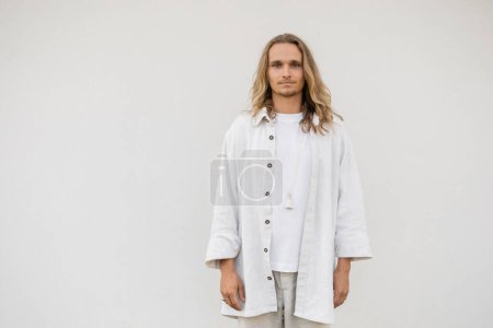 stylish yoga man with long fair hair standing near white wall and looking at camera