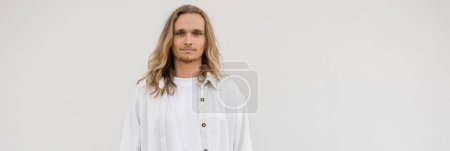 retrato de joven hombre de yoga de pelo largo en camisa de lino mirando a la cámara cerca de la pared blanca, pancarta