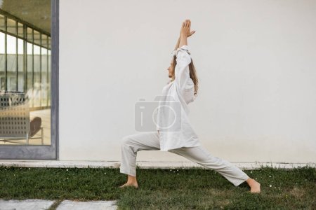 Téléchargez les photos : Vue latérale de l'homme pieds nus dans des vêtements de coton blanc méditant dans la posture guerrière avec les mains levées priant près du chalet moderne - en image libre de droit