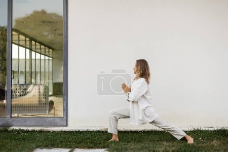 Foto de Vista lateral del hombre de pelo largo en ropa de lino meditando en pose guerrera con las manos de oración cerca de la pared blanca de la casa de campo moderna - Imagen libre de derechos