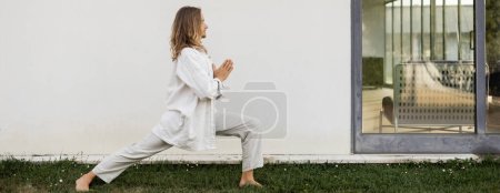 Téléchargez les photos : Vue latérale de l'homme élégant en vêtements blancs pratiquant le yoga en posture guerrière avec geste anjali mudra près de la maison moderne, bannière - en image libre de droit