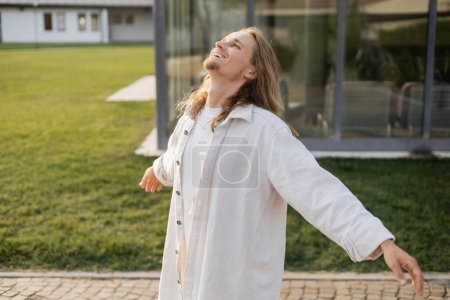 carefree yoga man in white linen shirt standing with outstretched hands and looking up near blurred house 