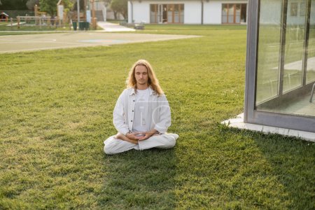 junger und stilvoller Mann in Leinenkleidung sitzt auf dem Rasen in der Nähe von Hütten und meditiert in Lotus-Pose