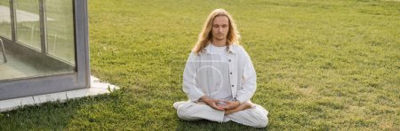 Photo for Young man in white clothes meditating in lotus pose on green lawn near building, banner - Royalty Free Image