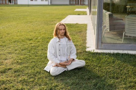 hombre de pelo largo con estilo meditando en pose de loto mientras está sentado en el césped cubierto de hierba cerca del edificio