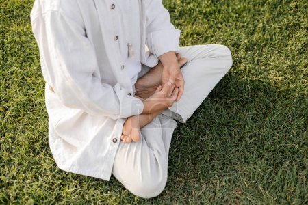 Teilansicht eines barfüßigen Mannes in weißen Leinenkleidern, der in Lotus-Pose auf grünem Rasen meditiert