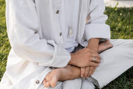 vista recortada del hombre en ropa blanca sentado en pose de loto durante la meditación al aire libre