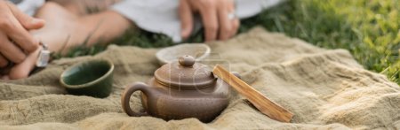 Photo for Partial view of blurred man sitting on green lawn near linen rug with clay teapot and cups, banner - Royalty Free Image