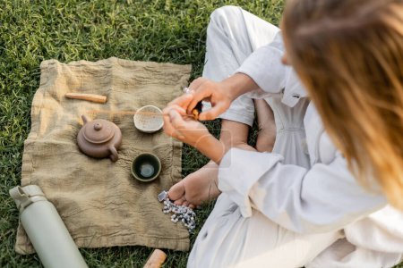 Téléchargez les photos : Vue du dessus de l'homme de yoga tenant bâton parfumé près tapis de lin avec théière d'argile et bols sur pelouse herbeuse - en image libre de droit
