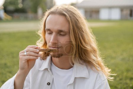 Téléchargez les photos : Homme de yoga aux cheveux longs avec les yeux fermés appréciant la saveur de palo santo bâton à l'extérieur - en image libre de droit