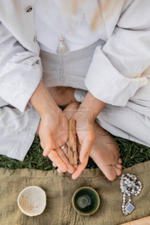 top view of cropped man in white clothes holding palo santo stick near linen rug with ceramic cups and mala beads 