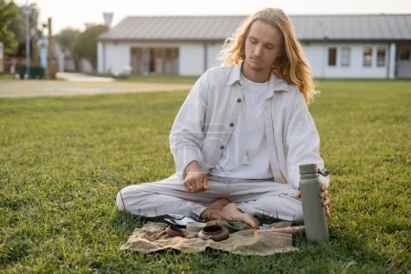 long haired man in white linen clothes holding thermos near ceramic teapot and bowls during tea ceremony in countryside