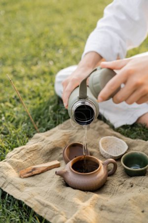 abgeschnittene Ansicht von Yoga-Mann, der heißes Wasser in Keramik-Teekanne gießt, während er auf Rasen in der Nähe von Leinenteppich mit Tassen und Palo Santo Stick sitzt