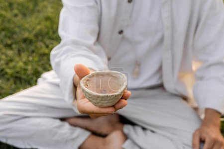 Teilansicht eines Yoga-Mannes in weißer Kleidung, der eine Tasse mit Tee im Freien hält