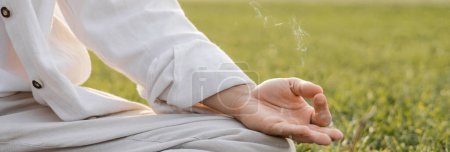 Téléchargez les photos : Vue recadrée de l'homme en lin montrant geste mudra gyan tout en méditant près de fumée parfumée sur la pelouse verte, bannière - en image libre de droit