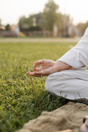 Foto de Vista recortada del hombre en ropa de lino blanco meditando con gesto de gyan mudra mientras está sentado en el campo de hierba verde - Imagen libre de derechos