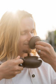 pleased long haired man with closed eyes holding oriental teapot and enjoying flavor of puer tea in sunshine outdoors t-shirt #648523180