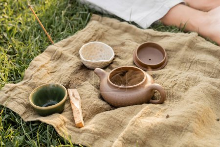 aromatischer Palo Santo Stick und Keramik-Teekanne mit Tassen in der Nähe von beschnittenen Yoga-Mann sitzt auf Rasen