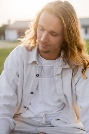 joven y soñador hombre con el pelo largo y claro meditando mientras está sentado al aire libre