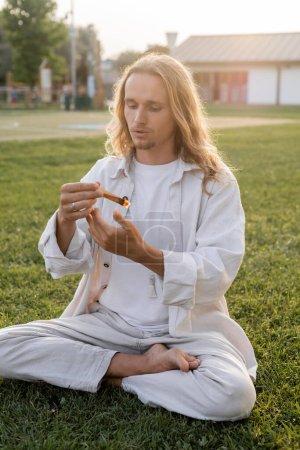 young long haired man in white linen clothes sitting in easy pose and holding scented palo santo stick on green lawn