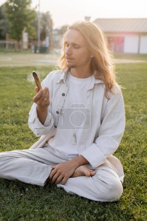 stilvoller Mann in weißen Leinenkleidern sitzt in leichter Pose und hält schwelenden Palo Santo Stock, während er im Freien meditiert
