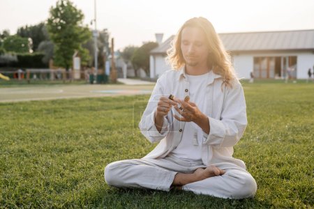Foto de Hombre de pelo largo en ropa de lino blanco sentado en pose de yoga fácil y sosteniendo palo santo más ligero y aromático al aire libre - Imagen libre de derechos